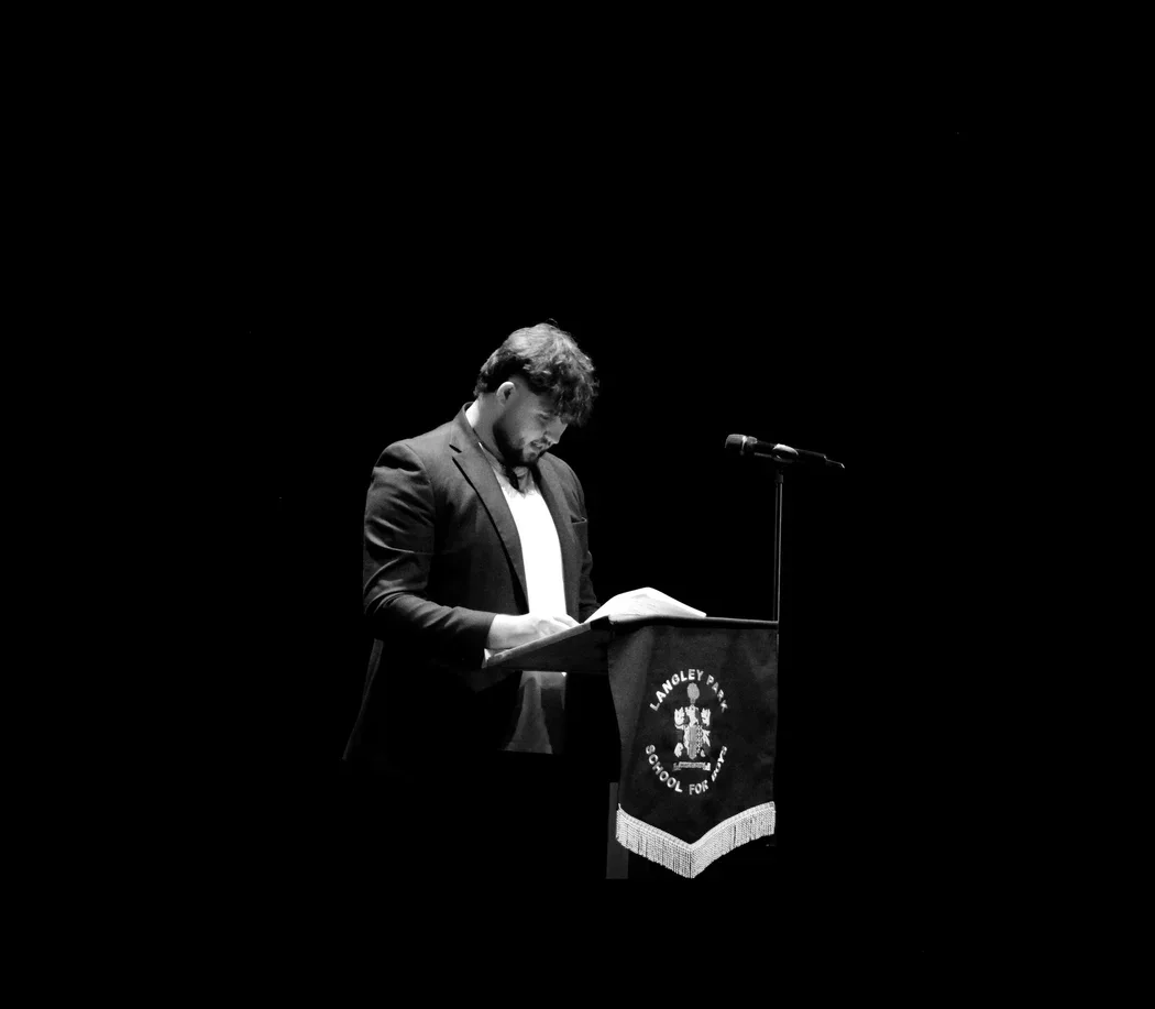 Brydon Duncan speaking at a lectern at Langley Park School, lit against a dark auditorium